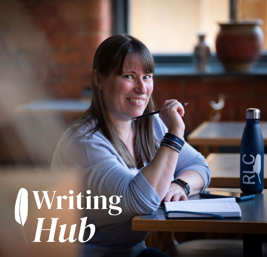 Writing Hub - Rebecca seated at a table wearing pale blue jumper with a book and pencil